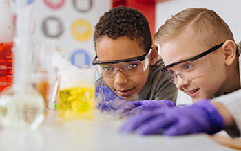 boys in safety goggles leaning closely to the table and watching chemical reaction going on in the beaker