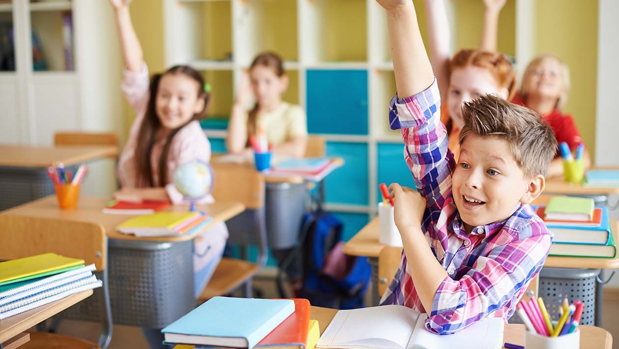 Boy in plaid shirt raising his hand in class with open notebook on his desk