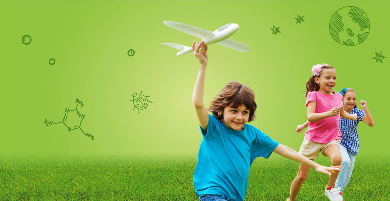 4 children running a green field with a tree in the background.  The child in front is holding a toy plane. 