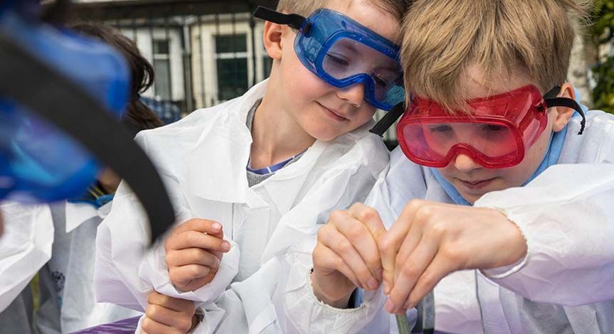 A girl and a boy wearing safety goggles and a mad science lab coat pouring liquid in a cup.