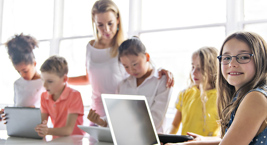 kids with tablets and laptop computer in classroom while teacher stands behind them
